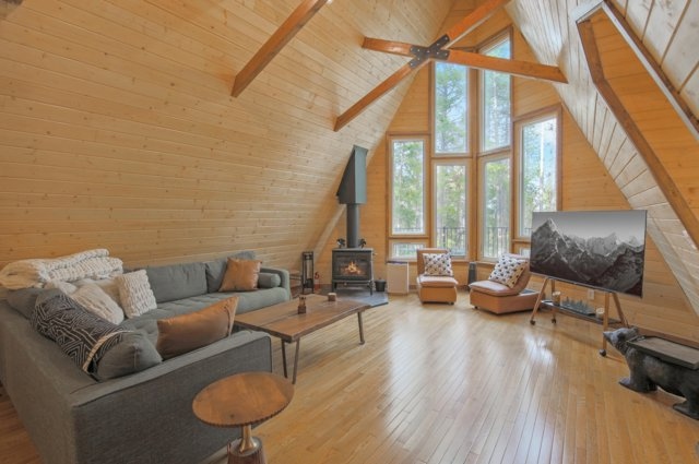 Vaulted A-frame great room at The Evergreen Aerie, with exposed timber beams, a wood stove, and floor-to-ceiling windows onto the pines